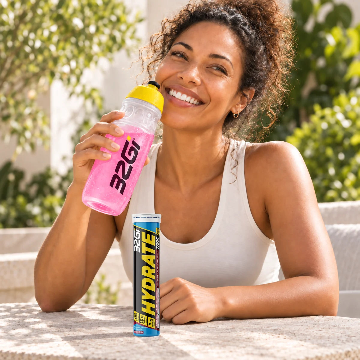 A woman outdoors, smiling, holding a 32GI bottle of Hydrate Raspberry with the tube on the table