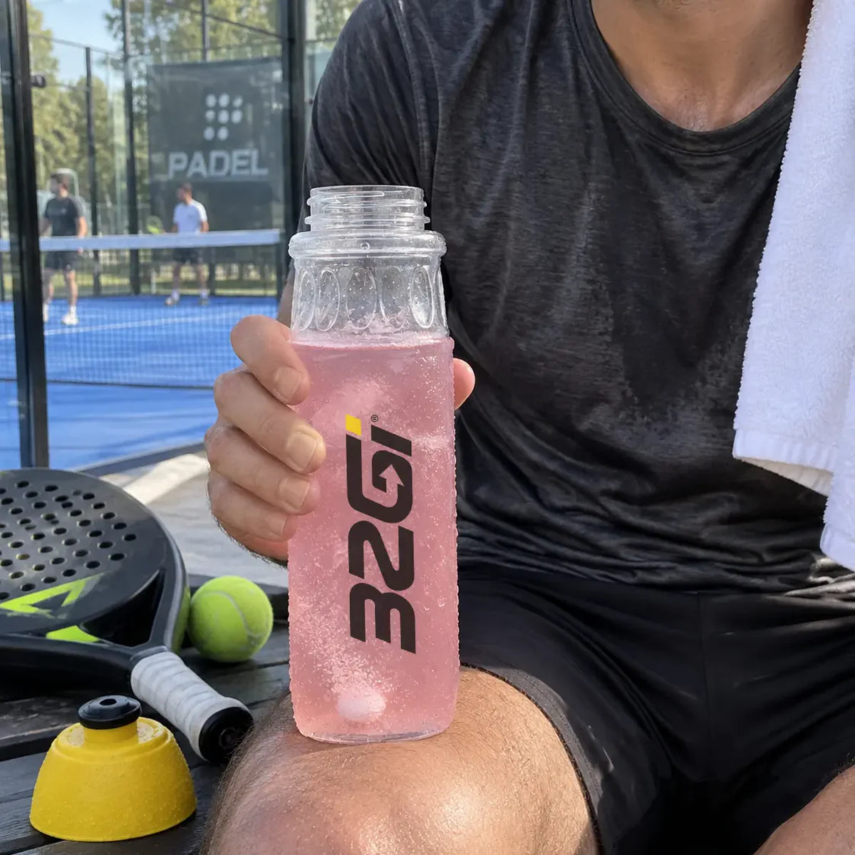 A padel player courtside with a towel and a 32GI bottle of Hydrate Raspberry, racket and balls at his feet