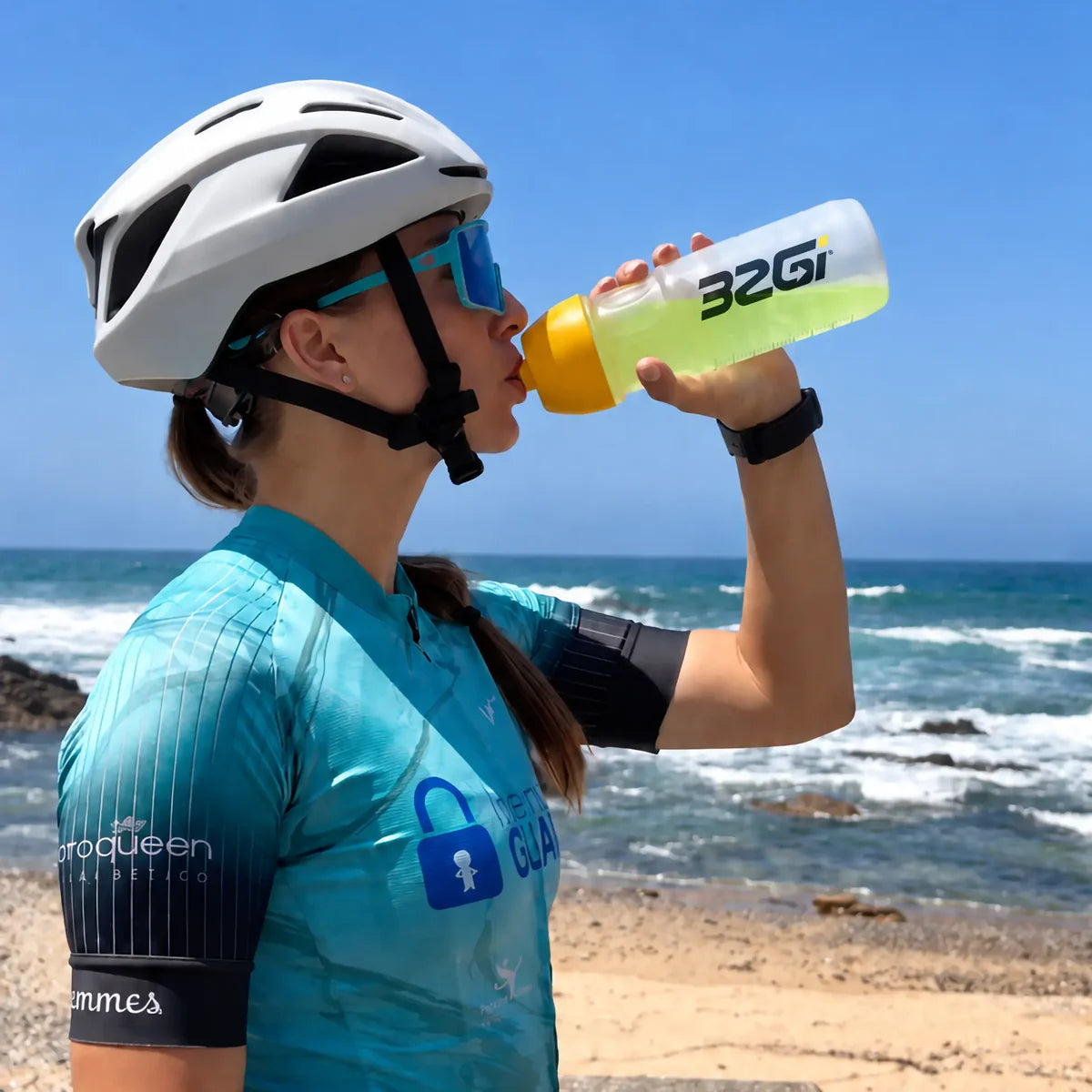 A cyclist in a helmet drinking green Hydrate from a 32GI bottle on a hot coastal road